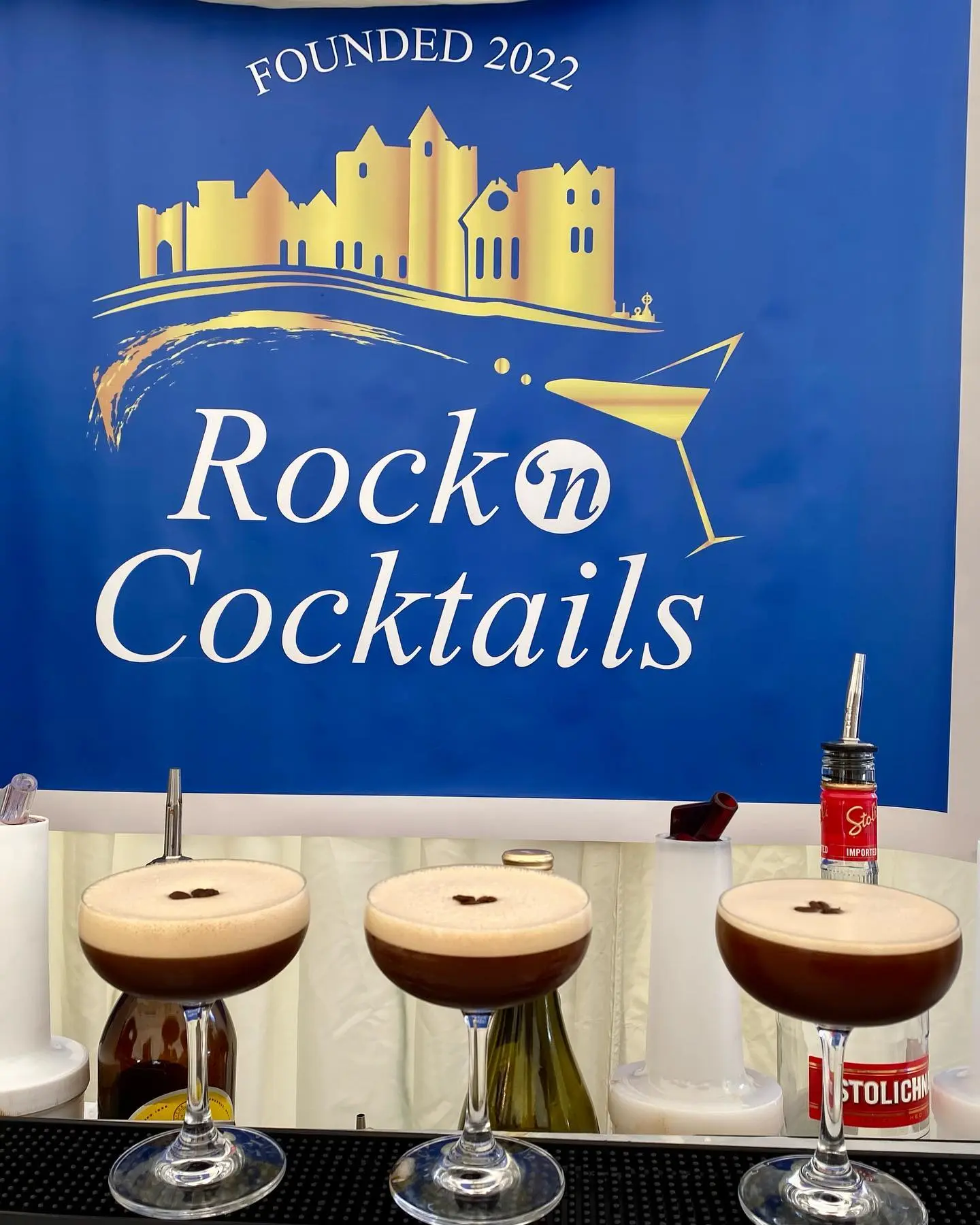 Three espresso martinis with foam and coffee bean garnish sit on a bar counter. Behind them is a blue banner with the &quot;Rock &lsquo;n Cocktails&quot; logo, gold illustrations of The Rock of Cashel, and the text &quot;Founded 2022&quot;.