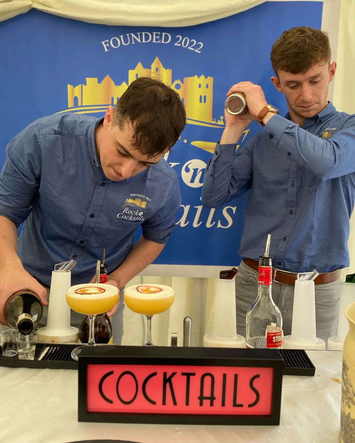 Two men in light blue shirts standing behind a sign that reads Cocktails. One is shaking a shaker, while the other is pouring from a bottle. On the bar are two passion fruit martinis in coupe glasses with a passion fruit garnish.