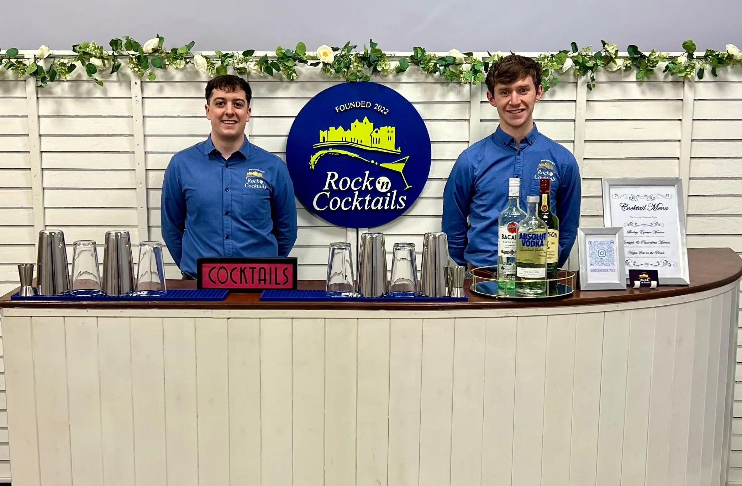 Two men in light blue shirts stand behind a bar with a cream front and a brown top. The bar top contains cocktail shakers, bottles of spirits, menus in picture frames and a cocktail sign. Behind the men is a cream wooden backdrop with a round blue sign with the "Rock ‘n Cocktails" logo, gold illustrations of The Rock of Cashel, and the text "Founded 2022".