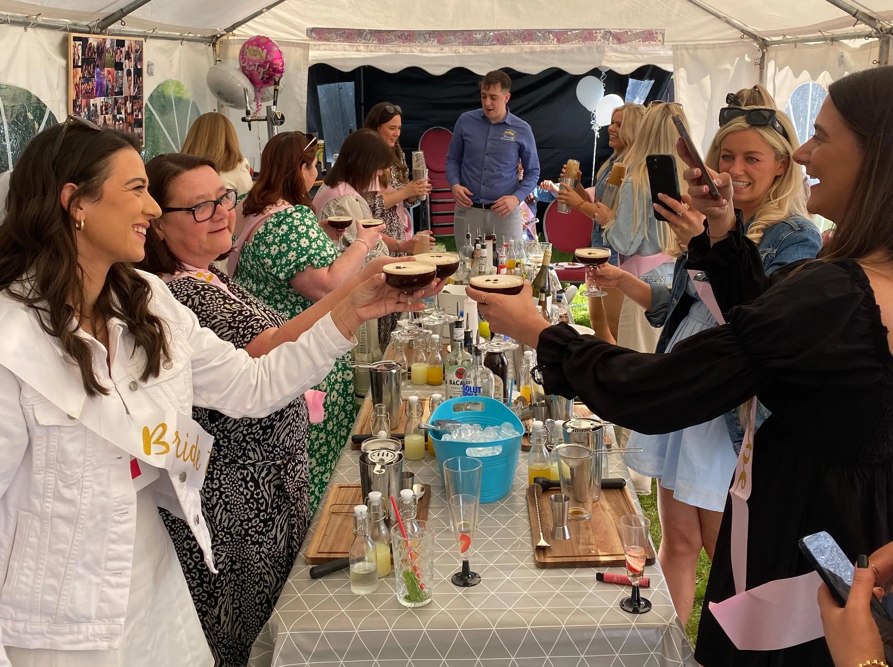 A group of woman cheers a series of espresso martini glasses while taking photos. The woman are standing around a table which contains cocktail making equipment.